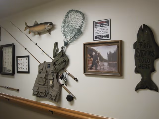 Wall decorated with fishing-themed items including a mounted fish, fishing rods, a fishing net, a fishing vest, framed pictures, and a wooden fish-shaped sign that reads 'Fish Stories Told Here'.