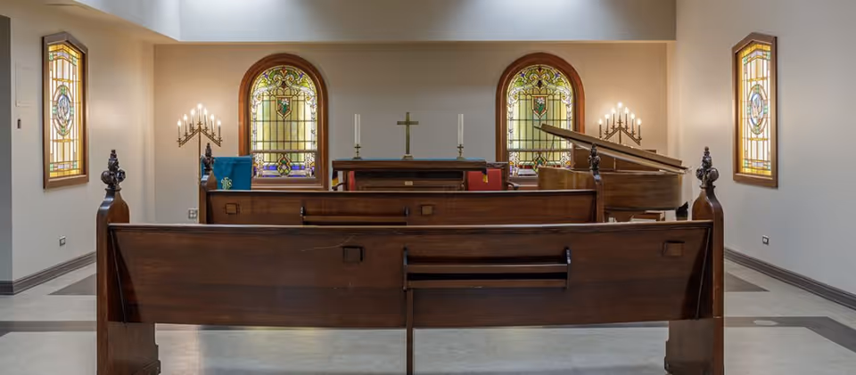Interior view of a small chapel or worship room with wooden pews, stained glass windows, a cross on the altar, two candle holders with lit candles, and a grand piano to the right side.