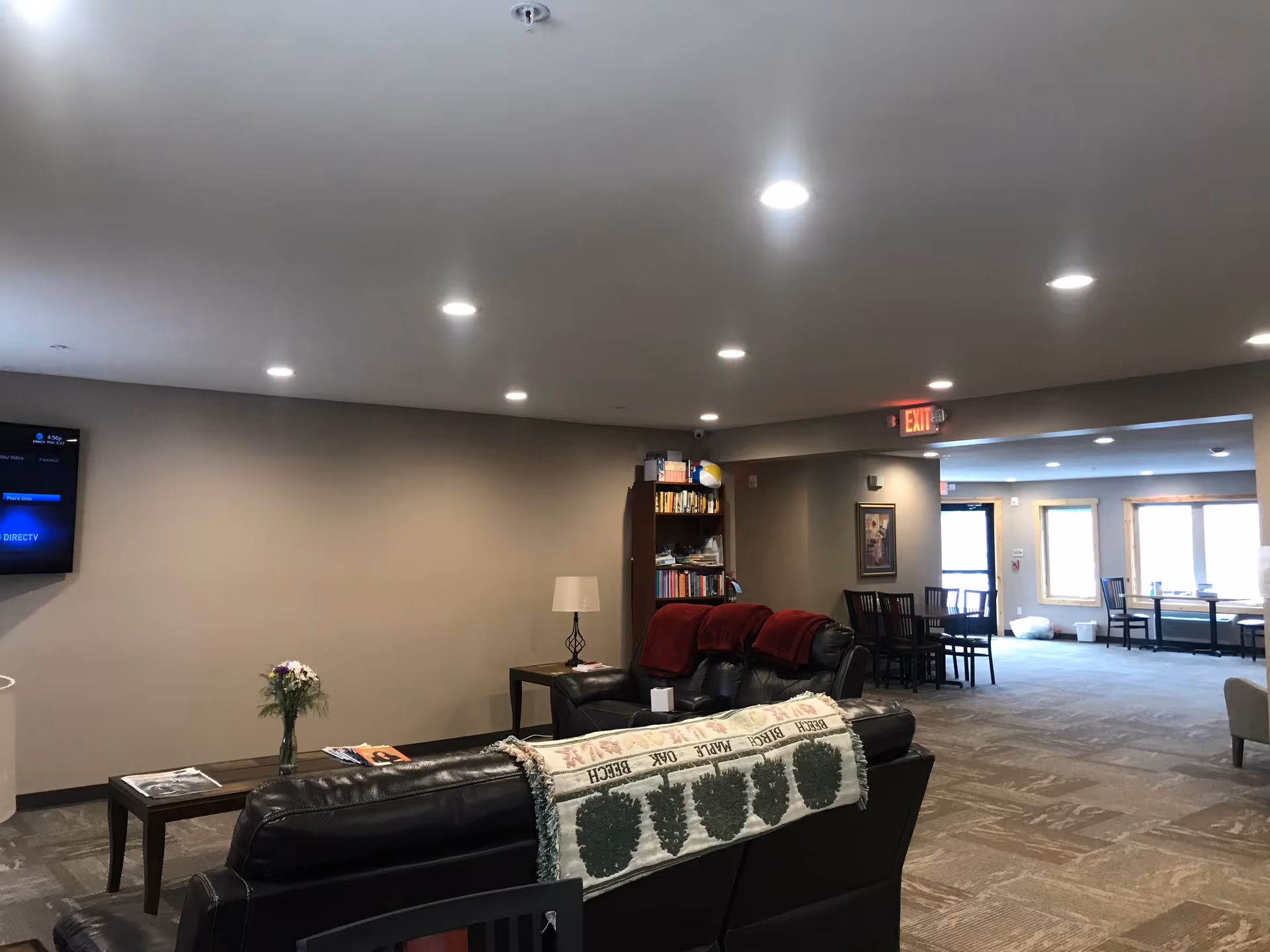 Spacious assisted-living common room with leather sofas, coffee tables, a bookshelf, wall-mounted TV, and a dining/seating area in the background.