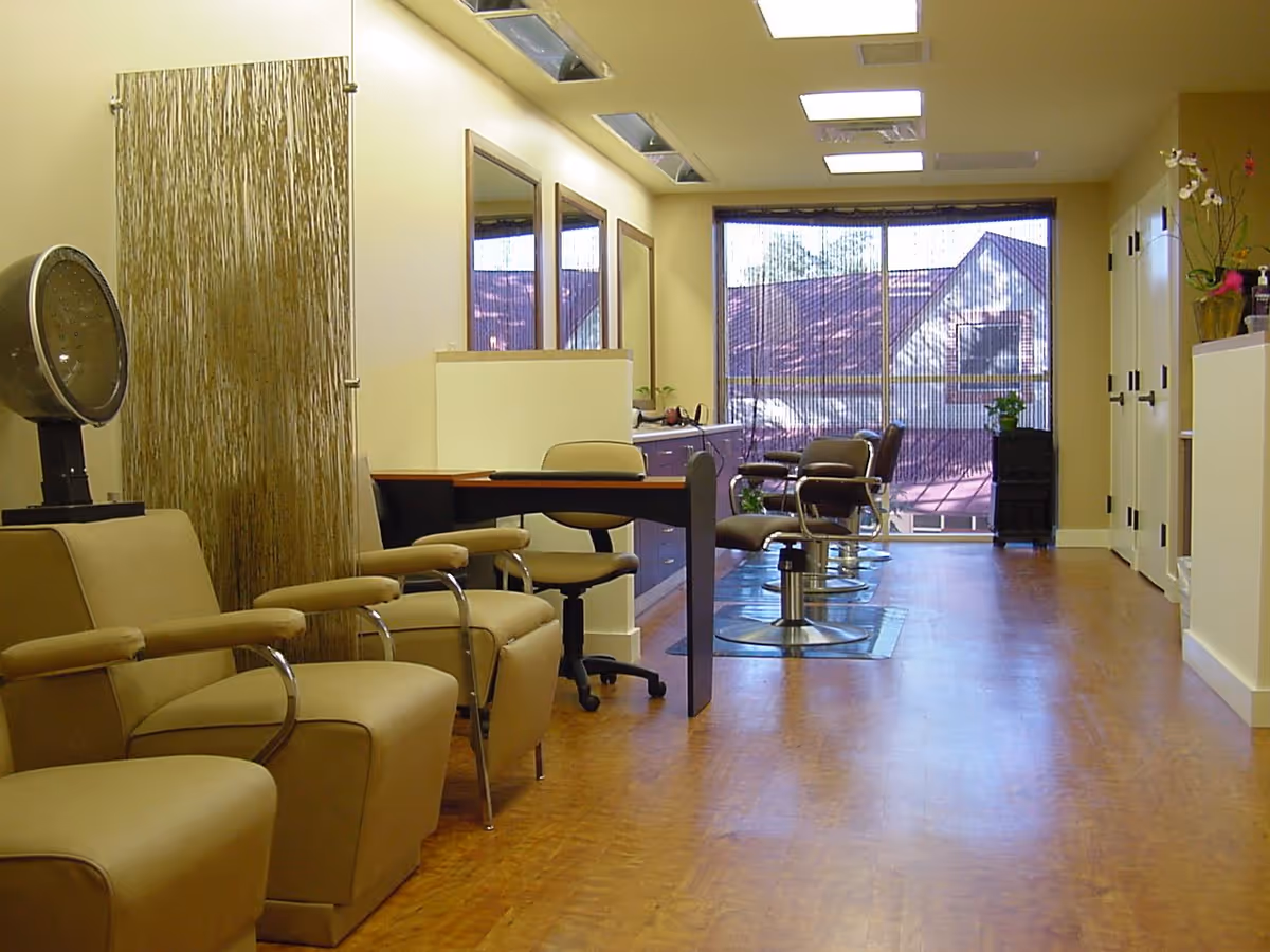 Interior view of a salon area in a senior living facility with beige salon chairs, a hair dryer, mirrors on the wall, a desk with a rolling chair, and large windows with a view outside.