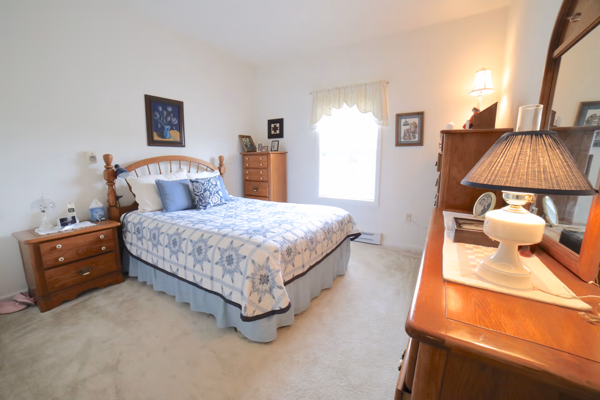 Sunlit bedroom with a wooden bed dressed in a blue patterned quilt, matching nightstands and dressers, and lamps.