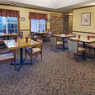 Dining room with several tables and chairs arranged neatly. Each table has a red cup and placemats. The room has carpeted flooring, two windows with red valances, and a brick accent wall.
