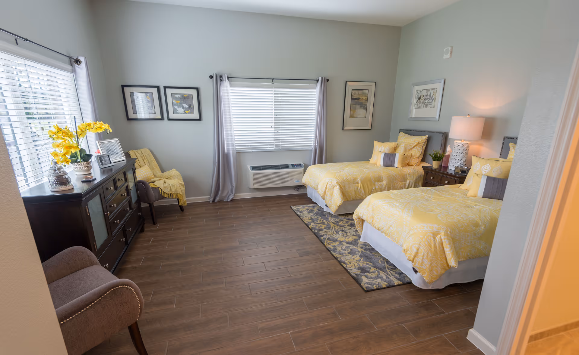 A bright and cozy bedroom with two twin beds featuring yellow bedding and decorative pillows. Between the beds is a nightstand with a lamp and small plant. The room has wood flooring, a patterned rug under the beds, and light gray walls adorned with framed artwork. A window with blinds and sheer curtains lets in natural light. On the left side, there is a dark wooden dresser with decorative items and a chair with a yellow throw blanket.