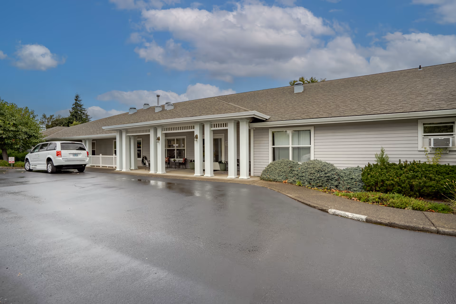 Front entrance of a single-story senior living facility with a covered portico, a parked white van, and surrounding landscaping.