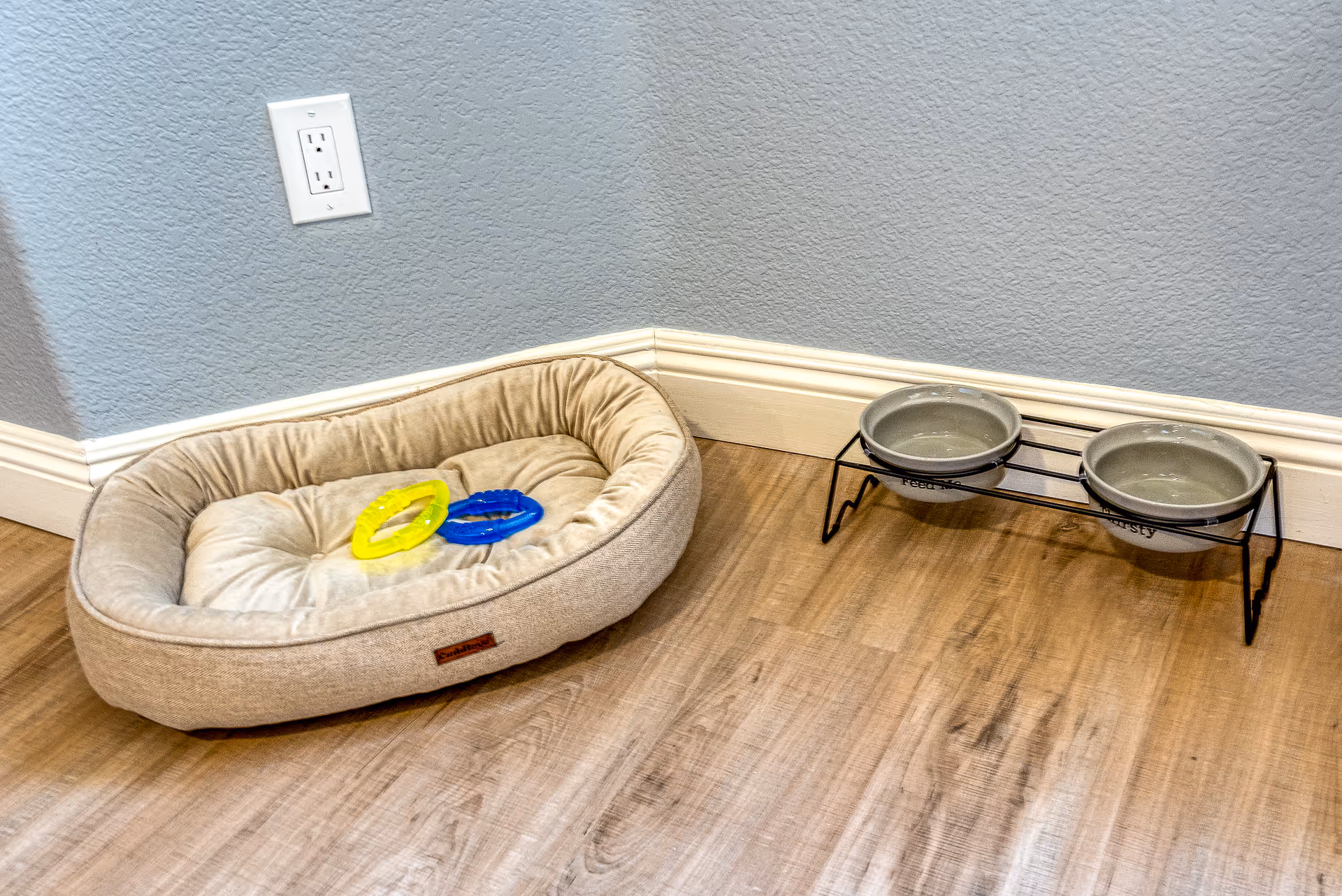 A beige pet bed with two colorful chew toys beside a raised double feeding bowl on a wood floor against a baseboard and electrical outlet.