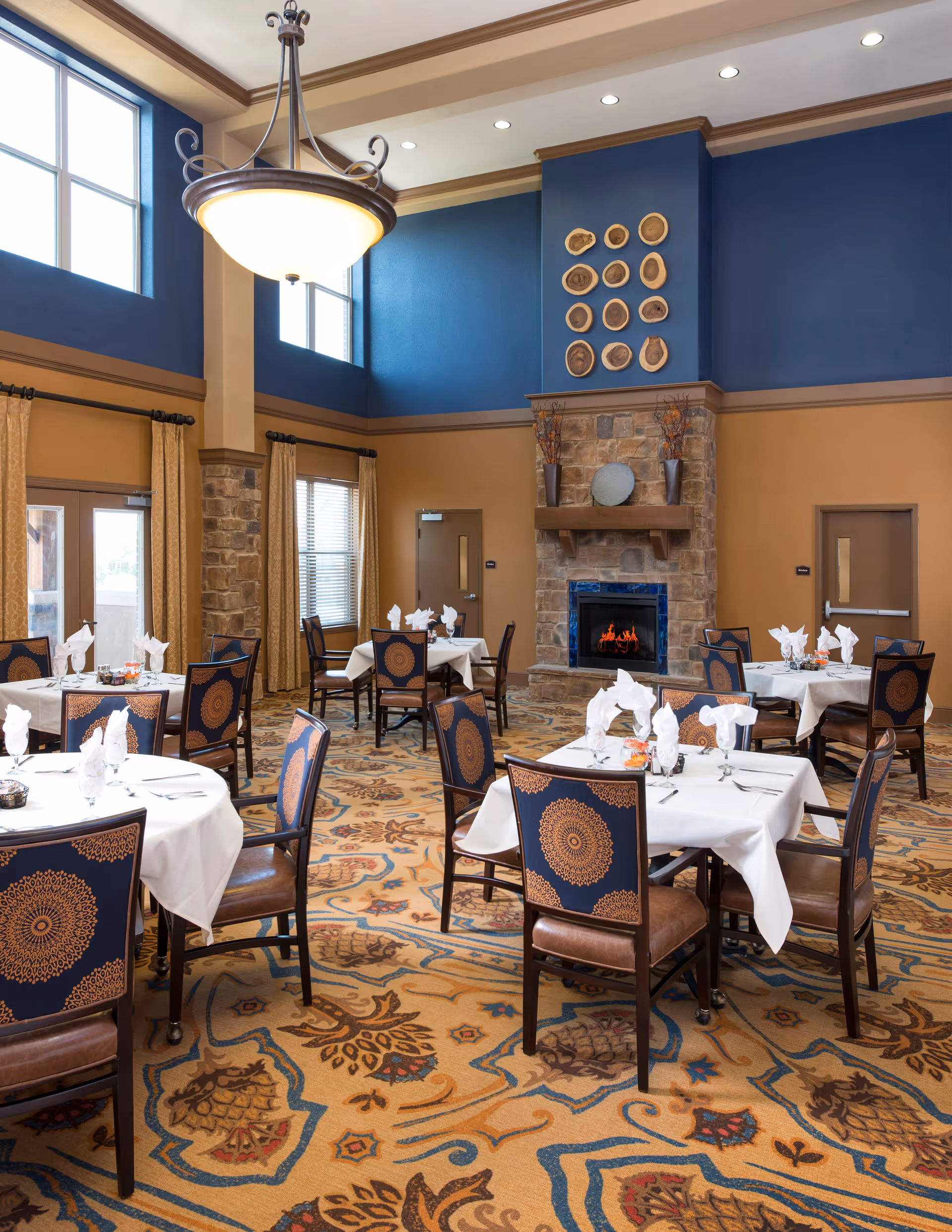 A dining room with several tables covered in white tablecloths, each set with napkins, glasses, and silverware. The room features a high ceiling with large windows, a decorative chandelier, and a stone fireplace with a fire burning. The walls are painted blue and tan, and the carpet has an intricate pattern. Chairs with blue and orange patterned upholstery surround the tables.