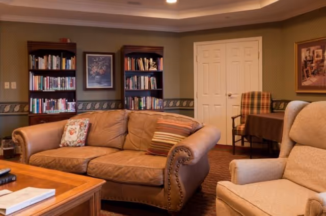 A cozy living room with a brown leather couch adorned with two decorative pillows, a beige armchair, two wooden bookshelves filled with books, a wooden coffee table with books on it, and a small table with a plaid chair in the background. The walls are decorated with framed artwork and have a green patterned wallpaper with a decorative border.