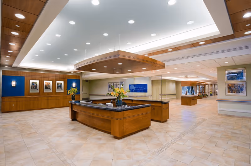 Spacious and well-lit reception area with a large wooden front desk adorned with flower vases. The ceiling features recessed lighting and a wooden accent structure above the desk. The walls have framed artwork and a blue accent panel behind the desk. The floor is tiled with a light beige pattern, and the hallway extends into the background with additional lighting and decor.