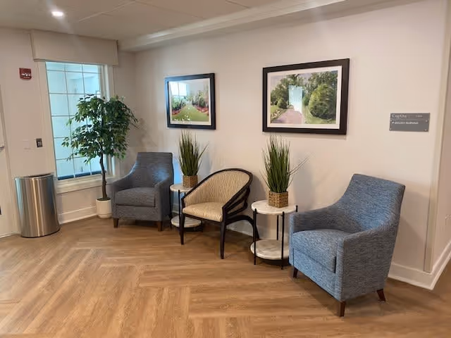 A cozy lobby area featuring two blue armchairs, a rattan chair, and decorative plants, with framed pictures on the wall.