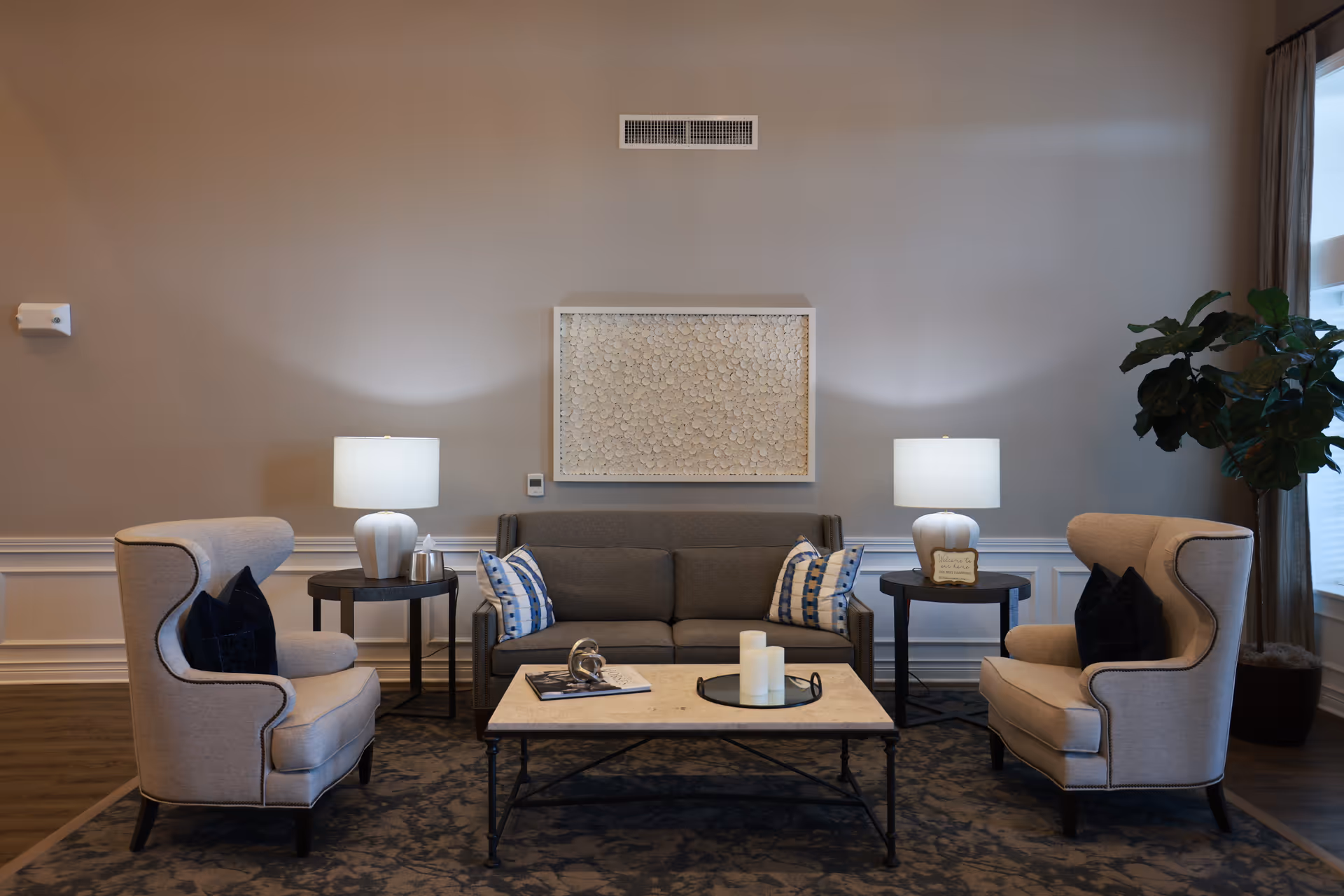 A cozy living room area with a gray sofa adorned with patterned pillows, flanked by two beige armchairs with dark cushions. Two side tables with white lamps are positioned on either side of the sofa. A rectangular coffee table with decorative items sits in front of the sofa on a patterned rug. A large potted plant is near a window with curtains, and a textured wall art piece hangs above the sofa.
