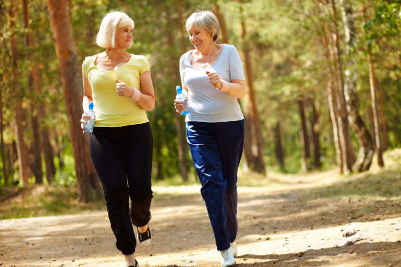 Two older women jogging on a forest trail, each holding a water bottle, surrounded by trees and sunlight filtering through the leaves.