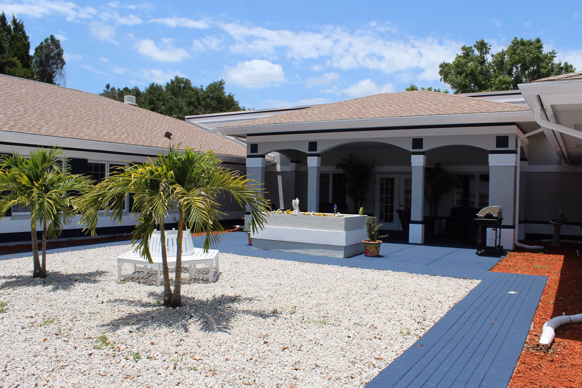 Outdoor courtyard area of Atrium Gardens Assisted Living with small palm trees, white gravel ground cover, a white circular bench around one tree, a raised rectangular planter, and a covered patio with seating under a blue sky with some clouds.