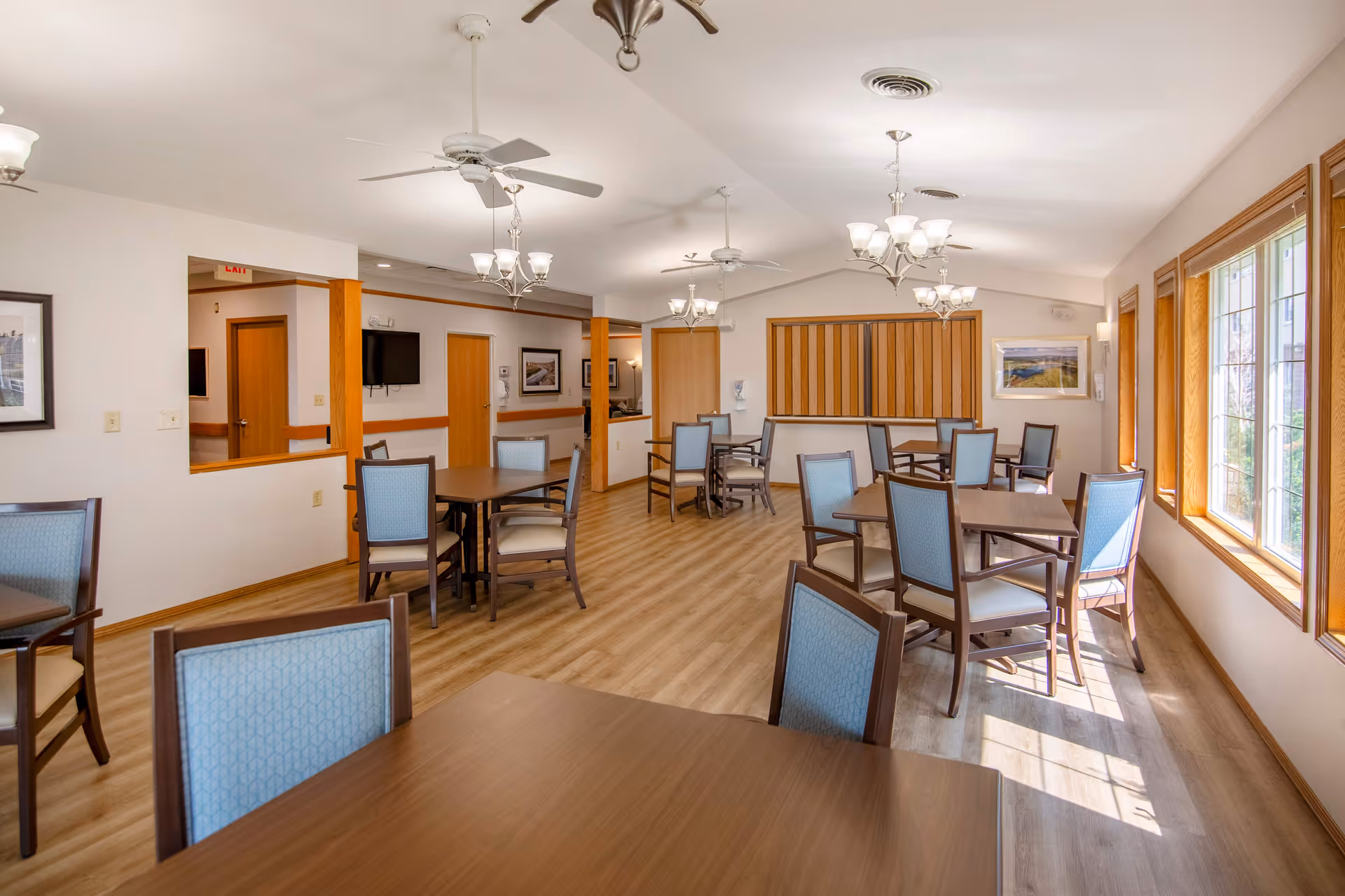 A bright and spacious dining room in a senior living facility with multiple wooden tables and chairs with blue upholstery. The room has large windows on one side letting in natural light, ceiling fans, and chandeliers providing additional lighting. The floor is wood, and there are framed pictures on the walls.