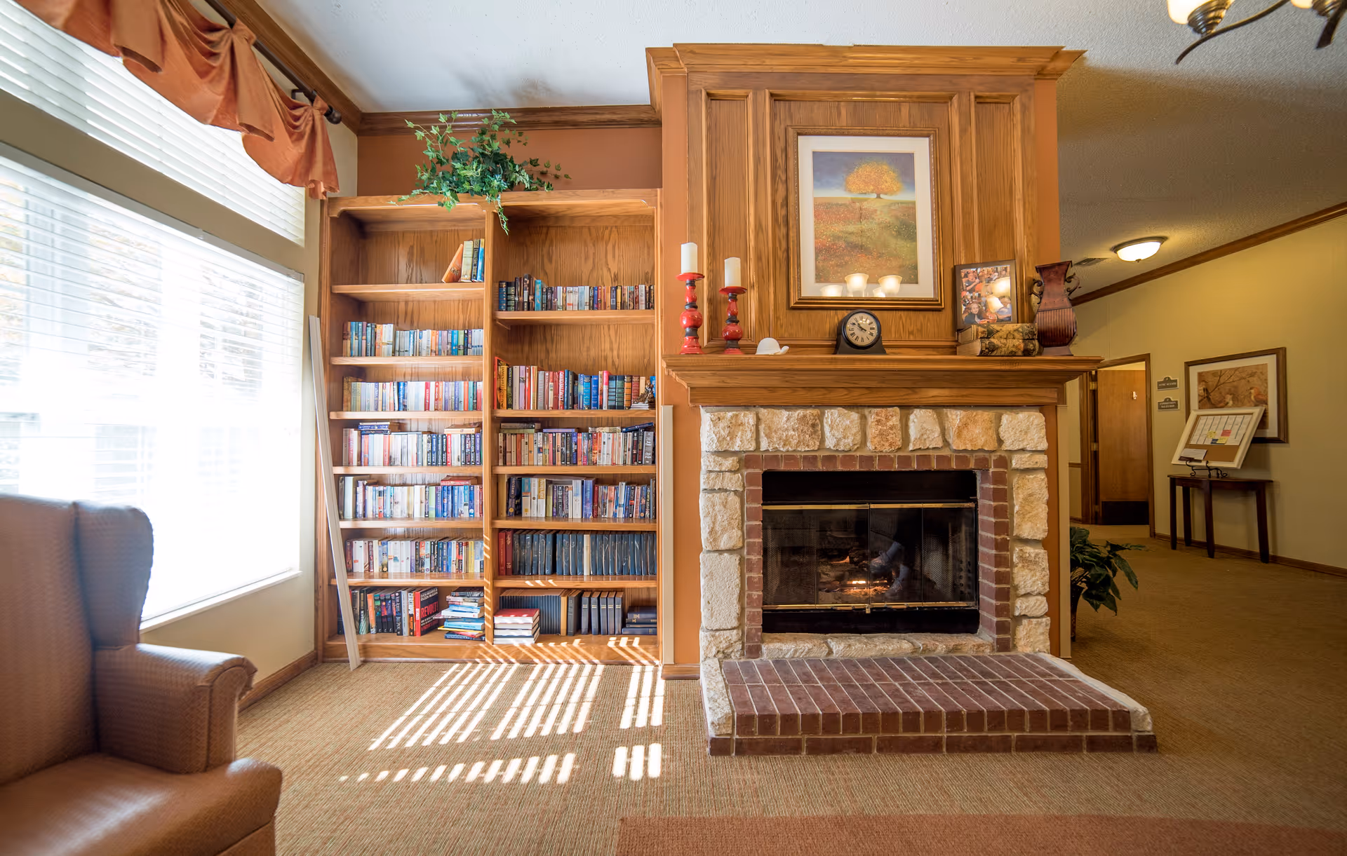 Cozy common room with a stone fireplace, built-in bookshelf, and armchair lit by a large window.