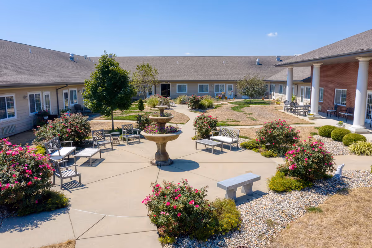 Outdoor courtyard area at Cedarhurst Senior Living of Highland featuring a central three-tiered fountain surrounded by benches and chairs. The courtyard is landscaped with flowering bushes, small trees, and neatly trimmed shrubs. The building surrounds the courtyard with multiple windows and doors, and there is a covered patio area with columns and additional seating.