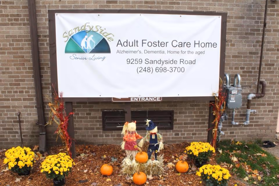Outdoor view of a brick wall with a large white sign for Sandyside Senior Living Adult Foster Care Home, including address and phone number. Below the sign are autumn decorations with two scarecrows, several small pumpkins, and yellow flowers arranged on the ground with fallen leaves.