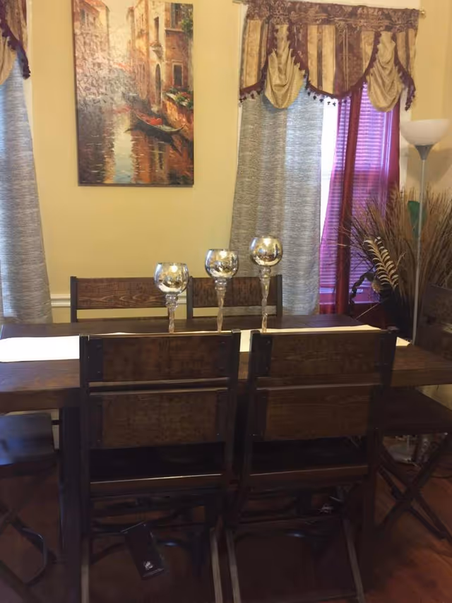 A dining room with a wooden table and six matching wooden chairs. On the table are three decorative silver candle holders. Behind the table, there are two windows with curtains and valances, and a floor lamp stands to the right. A painting depicting a canal scene with a gondola hangs on the wall.