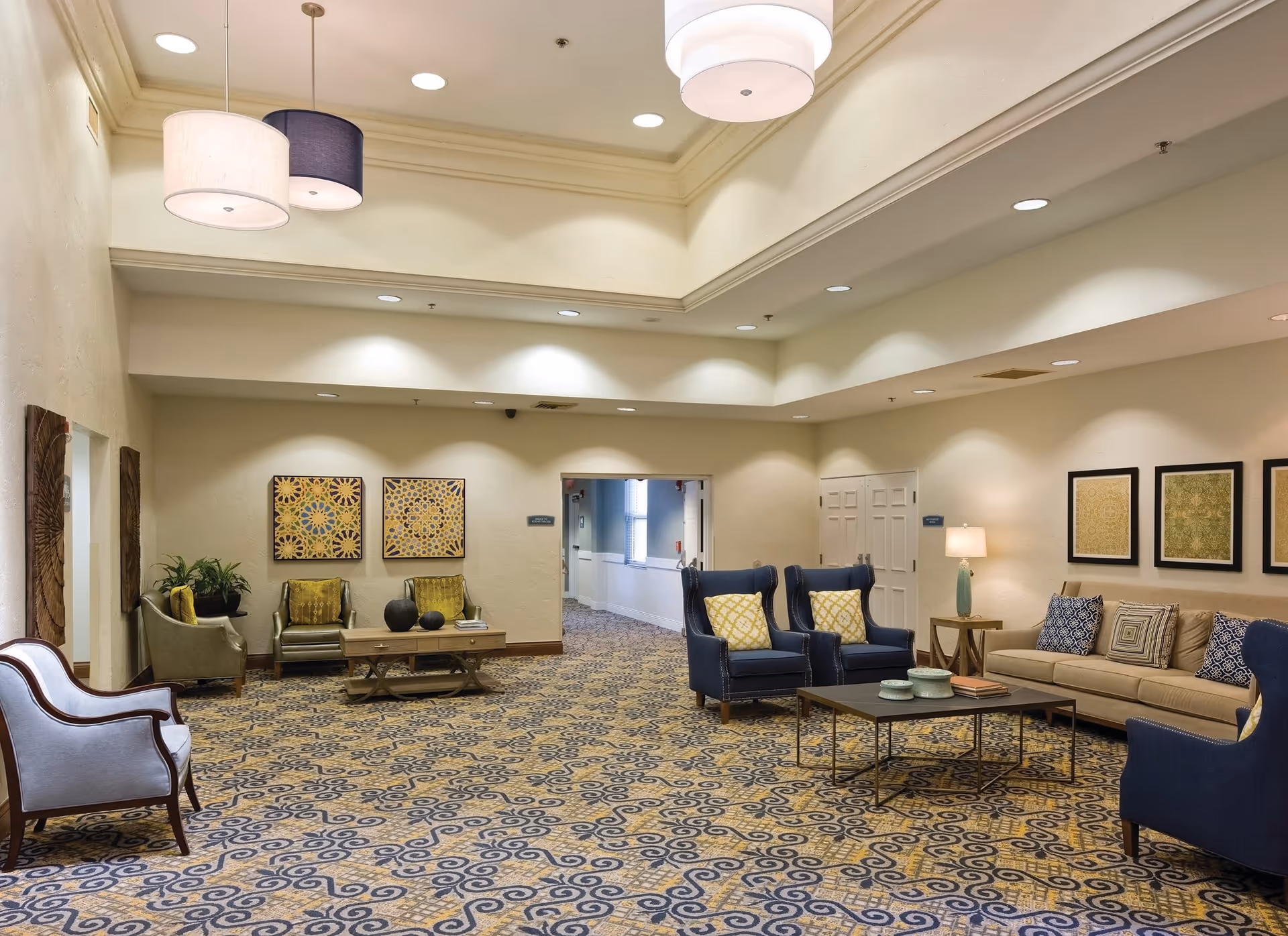 A spacious and well-lit common area in a senior living facility with patterned carpet, multiple seating arrangements including blue armchairs, a beige sofa with decorative pillows, and green chairs. The walls are adorned with framed artwork and there are hanging ceiling lights and recessed lighting.