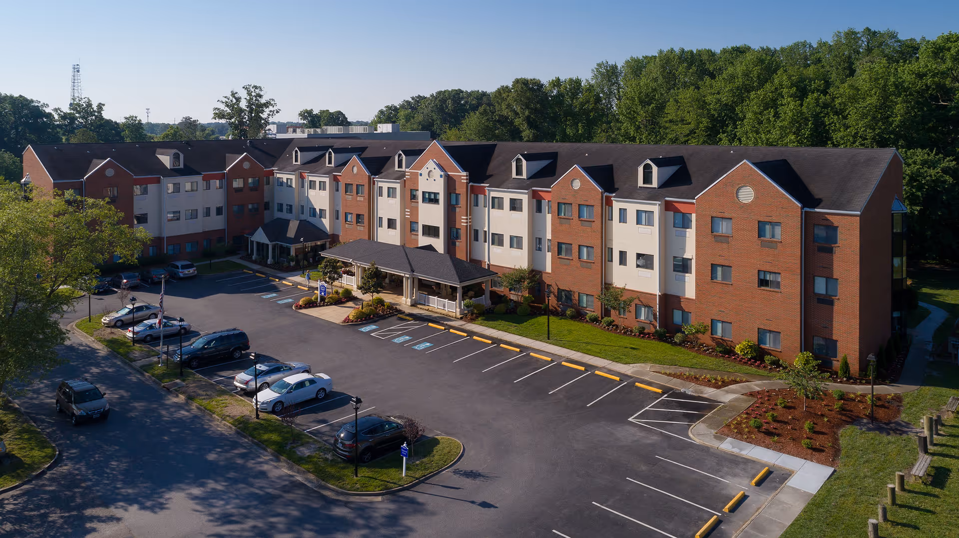 Aerial view of a three-story brick and beige panel building with a covered entrance and a parking lot in front, surrounded by trees and greenery under a clear blue sky.
