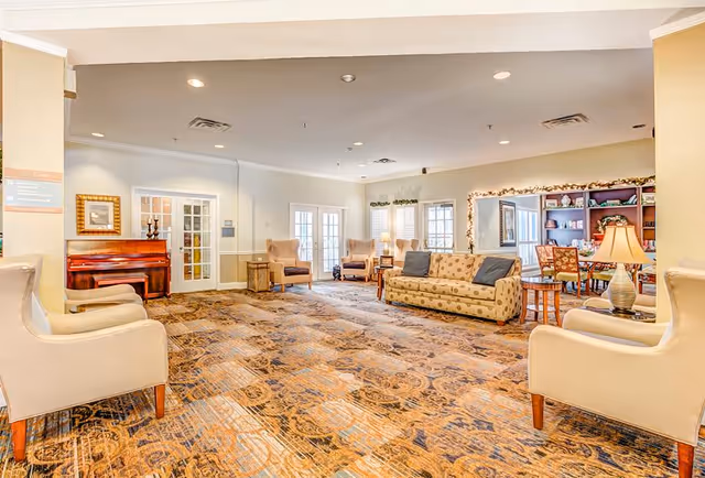 A spacious and well-lit living room area in a senior living facility with patterned carpet, beige walls, and recessed lighting. The room features multiple armchairs and a sofa with cushions arranged around small wooden tables. There is a piano against one wall, a large mirror with string lights, and a bookshelf filled with books and decorative items. French doors and windows allow natural light to enter the room.