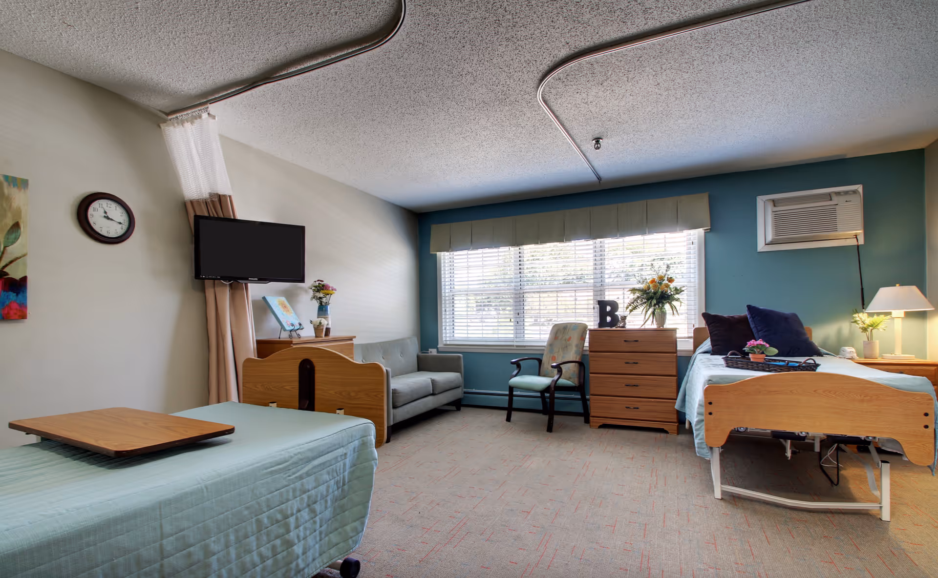 A well-lit senior living facility room with two hospital-style beds, a small sofa, an armchair, a chest of drawers, and a large window with blinds. The room has light blue and beige walls, a wall-mounted TV, a clock, and various decorative items including flowers and pillows.