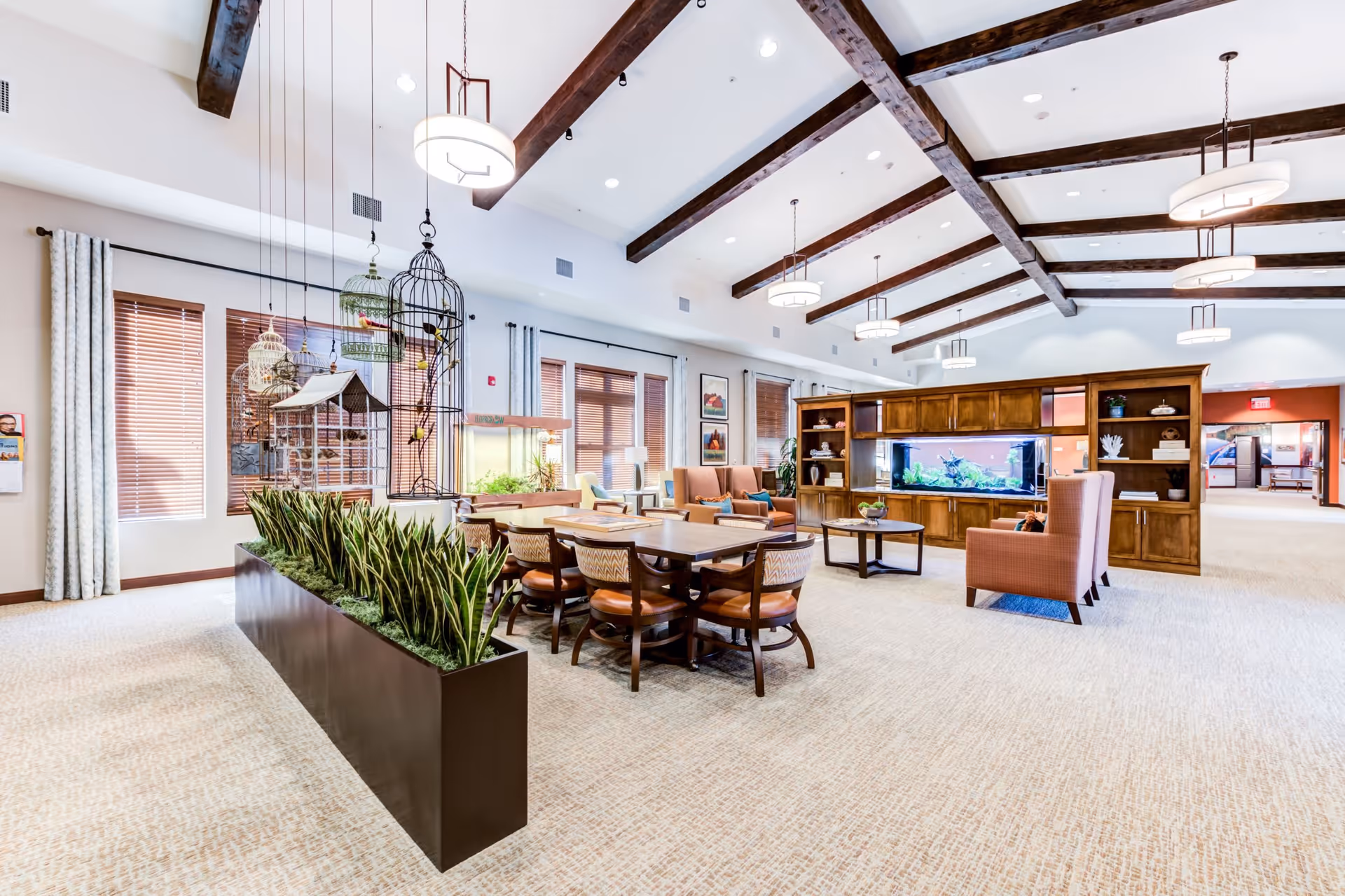 A spacious and well-lit senior living common area with high ceilings and exposed wooden beams. The room features a long table with chairs, a large planter with tall green plants, several birdcages hanging from the ceiling, a wooden cabinet with a built-in aquarium, comfortable armchairs, and large windows with wooden blinds and light curtains.