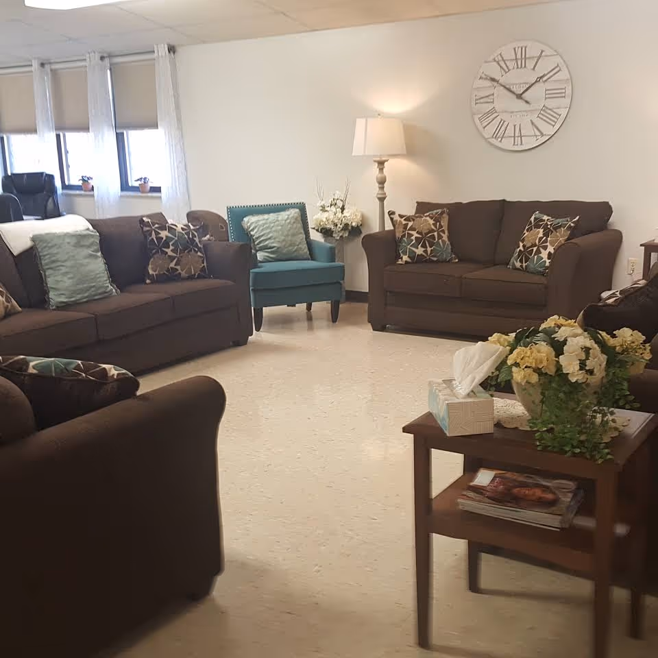 A bright communal living room with brown sofas, patterned throw pillows, a teal armchair, a floor lamp, a large wall clock and a side table with flowers and magazines.