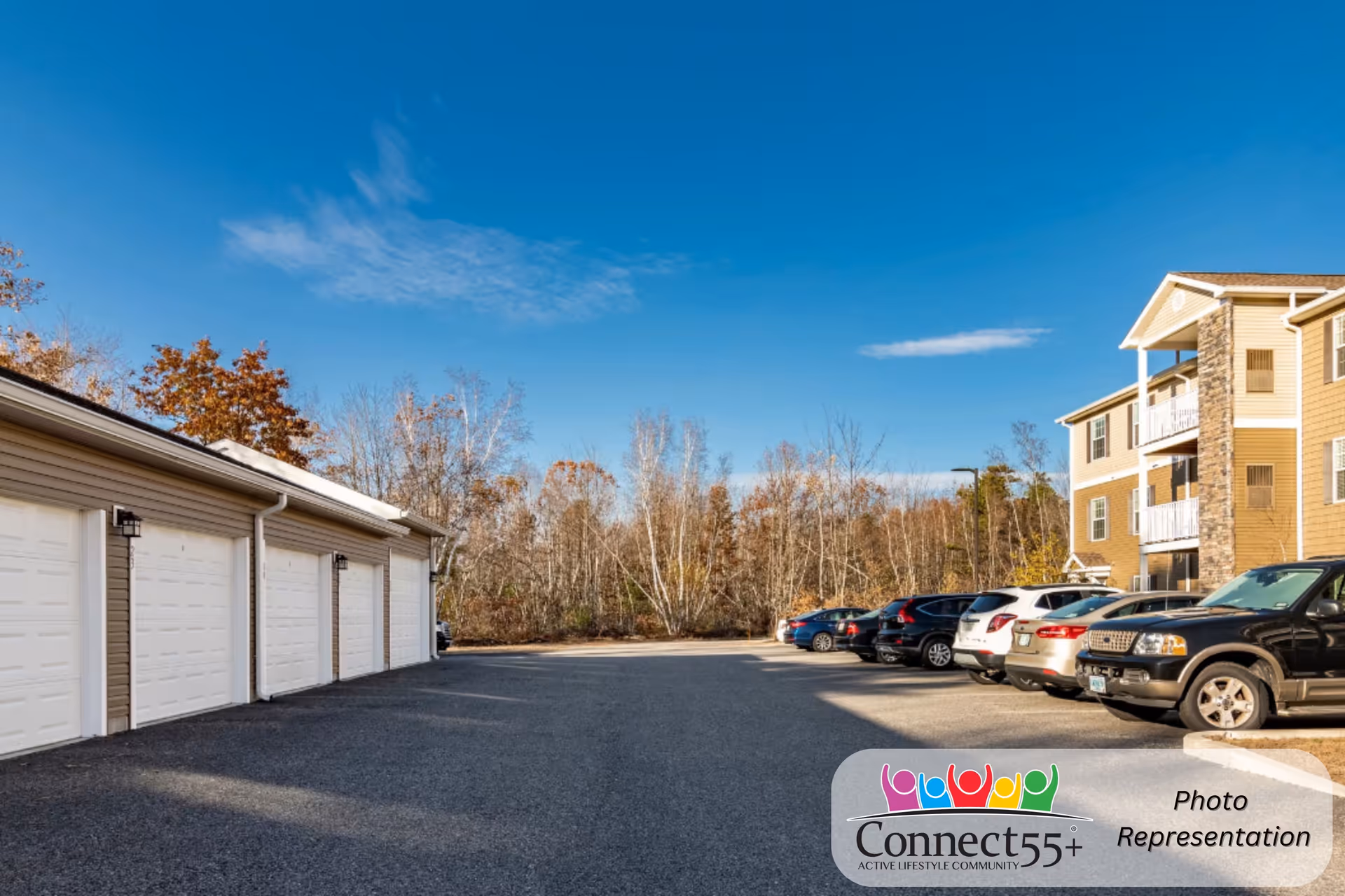 Parking area outside a senior living community with several parked cars and a row of garages on the left. A three-story building with balconies is visible on the right, and trees with autumn foliage are in the background under a clear blue sky.