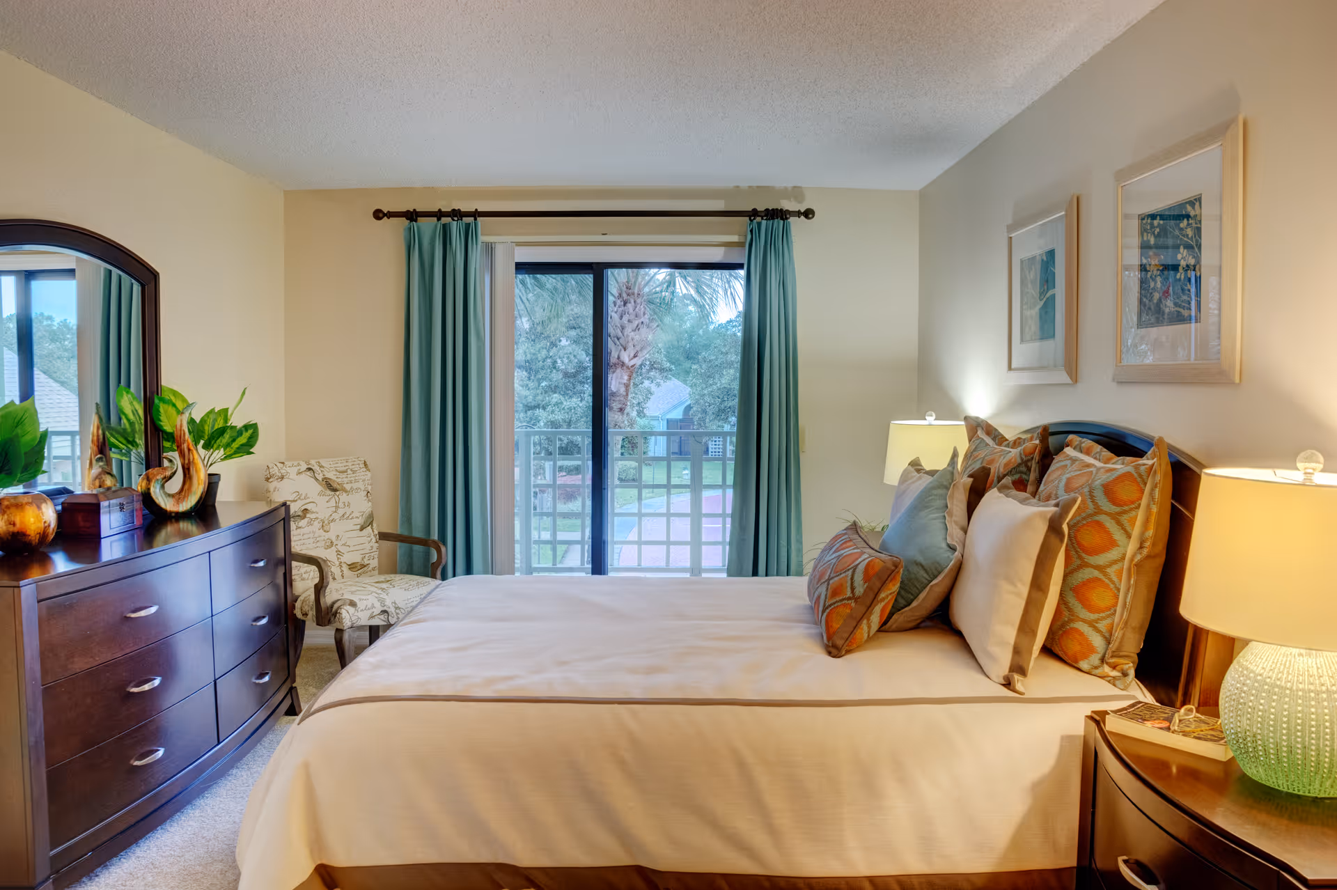 A cozy bedroom with a neatly made bed featuring multiple decorative pillows. There is a wooden dresser with a large mirror on the left side, adorned with plants and decorative items. A patterned armchair is placed near the sliding glass door, which is covered with blue curtains and opens to a view of trees and a building outside. Two framed artworks hang on the wall above the bed, and two bedside tables with lamps provide warm lighting.