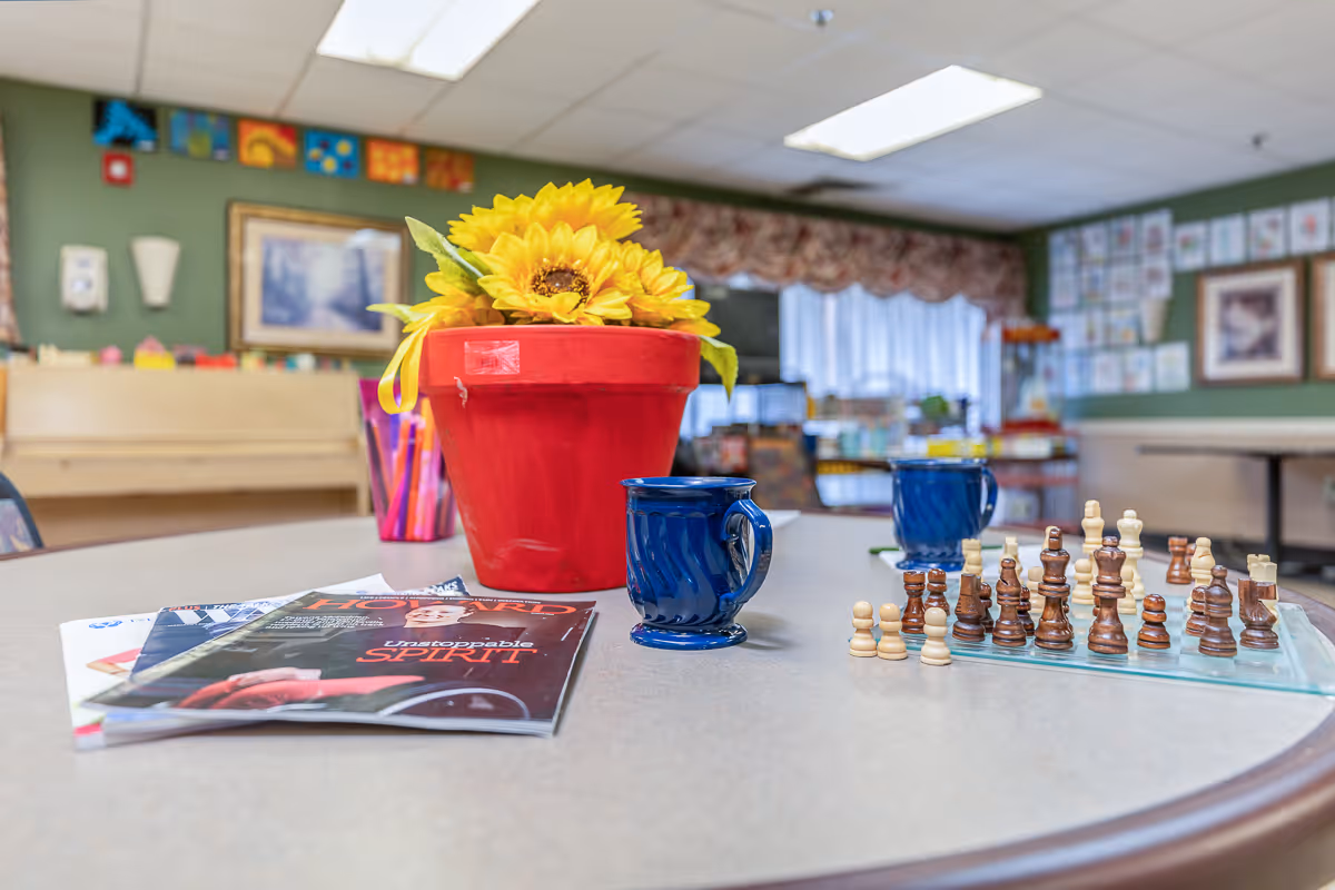 A communal activity table with a red pot of sunflowers, two blue mugs, magazines and a chess set, with a blurred activity room in the background.