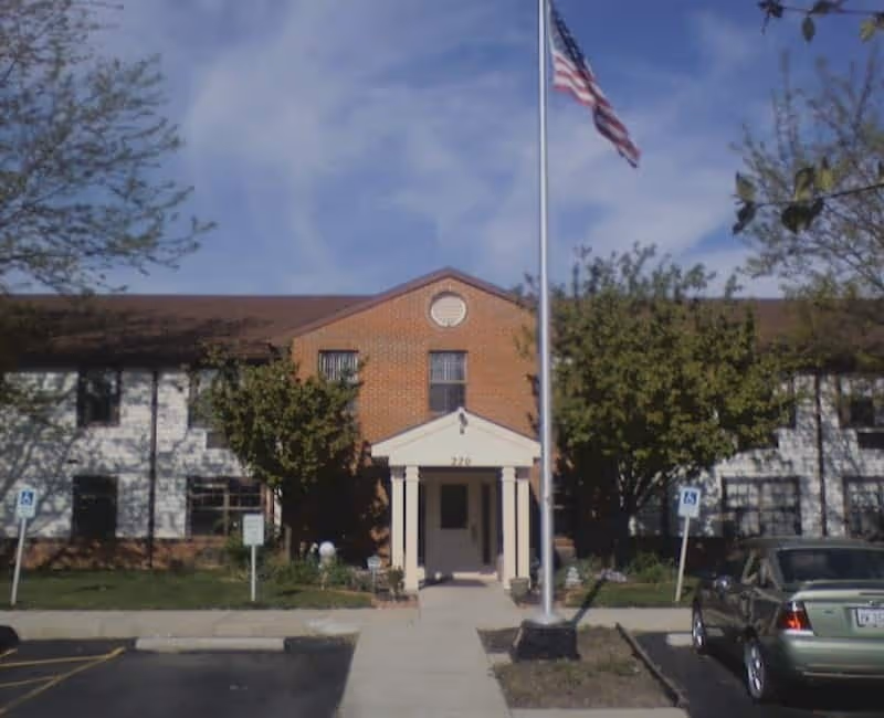 Front exterior view of a two-story brick and siding building with a covered entrance supported by columns, an American flag on a flagpole in front, trees on either side, and a parking lot with cars and handicap parking signs.