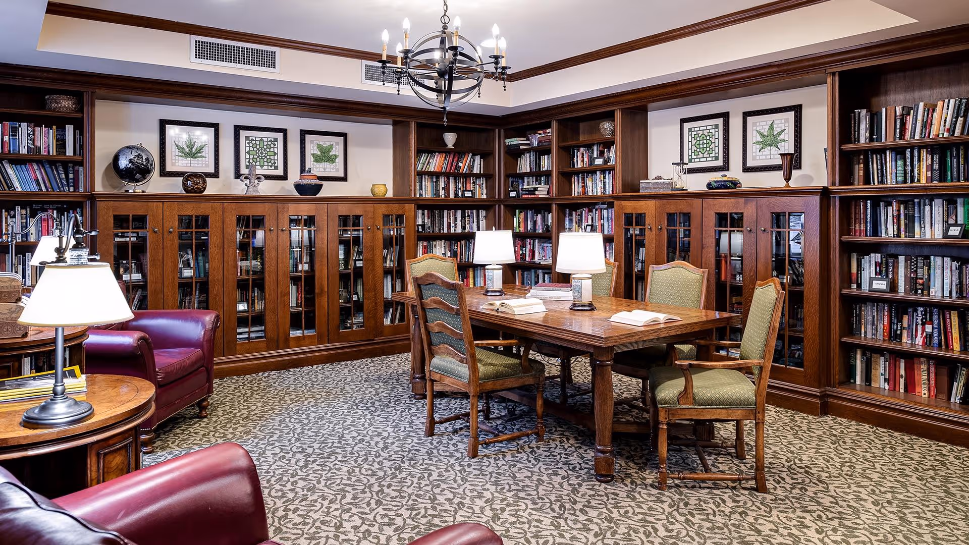 A cozy library room with wooden bookshelves filled with books lining the walls. In the center, there is a wooden table with four green upholstered chairs around it, and two table lamps providing light. Two red leather armchairs are positioned near a round wooden side table with a lamp. The room has a patterned carpet and a chandelier hanging from the ceiling. Framed botanical prints are displayed above the bookshelves.