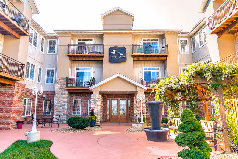 Entrance courtyard and front facade of The Fremont senior living building with balconies, a fountain, and seating.