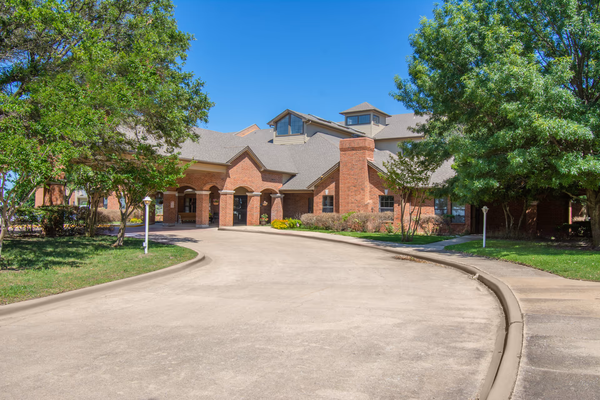 Exterior view of a brick building with a covered entrance and multiple roof peaks, surrounded by green trees and a clear blue sky.