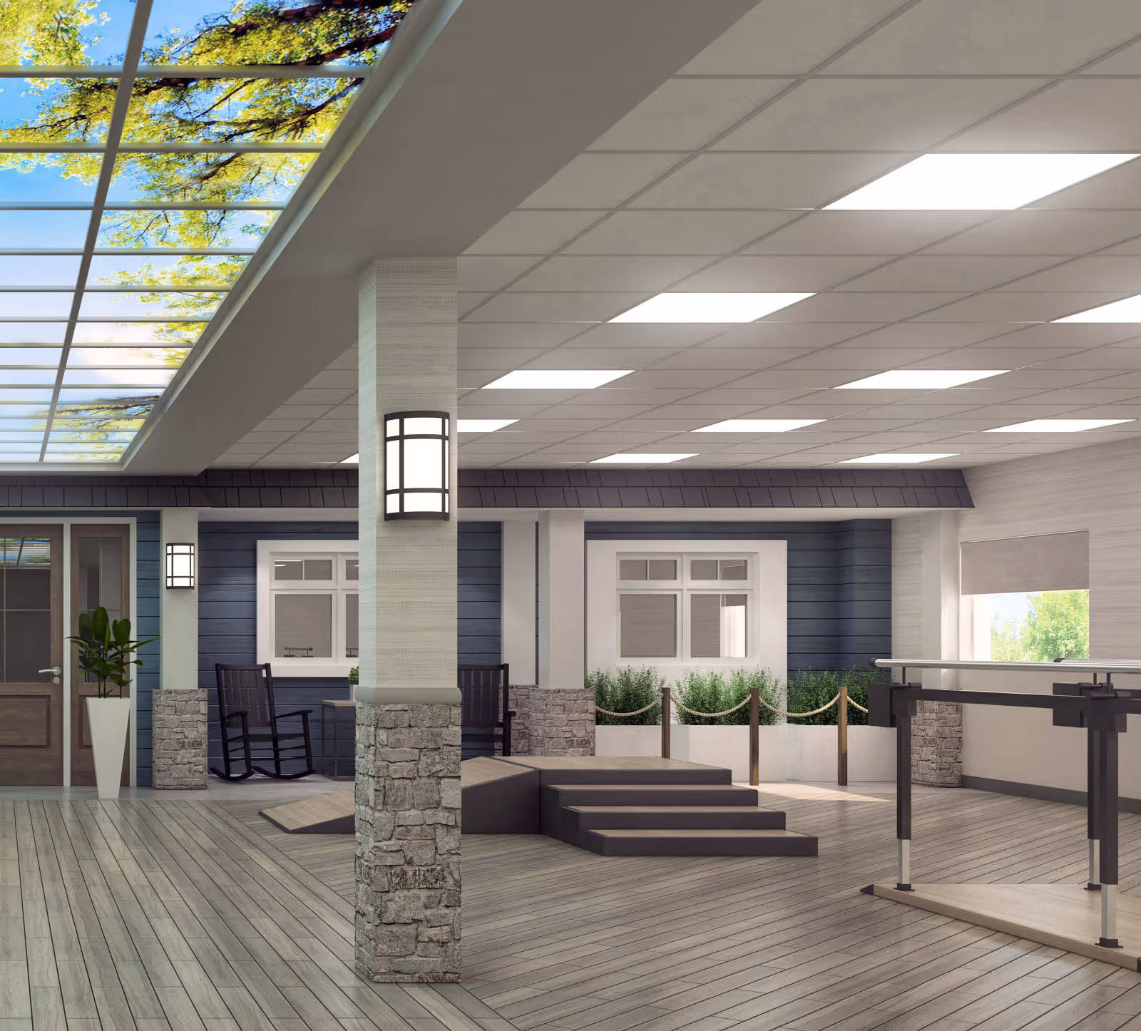 A spacious indoor common area with wooden flooring and a ceiling featuring bright rectangular lights and a skylight with a view of tree branches and blue sky. The room includes a stone pillar with a wall-mounted light, a small set of steps leading to a raised platform, a railing for support, and a seating area with rocking chairs near windows and a door. There are also plants and a rope barrier along the back wall.