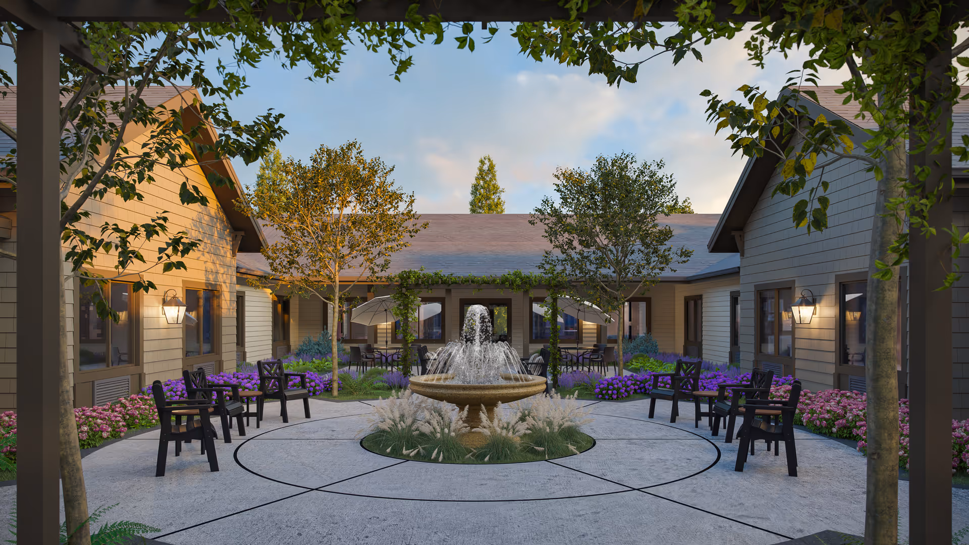 Outdoor courtyard area at Fields Senior Living at Spokane Valley featuring a central water fountain surrounded by a circular paved walkway. There are several black chairs arranged around the courtyard, with trees, flowering plants, and shrubs adding greenery. The courtyard is enclosed by single-story buildings with beige siding and windows, illuminated by wall-mounted lantern-style lights. The sky is partly cloudy with soft evening light.