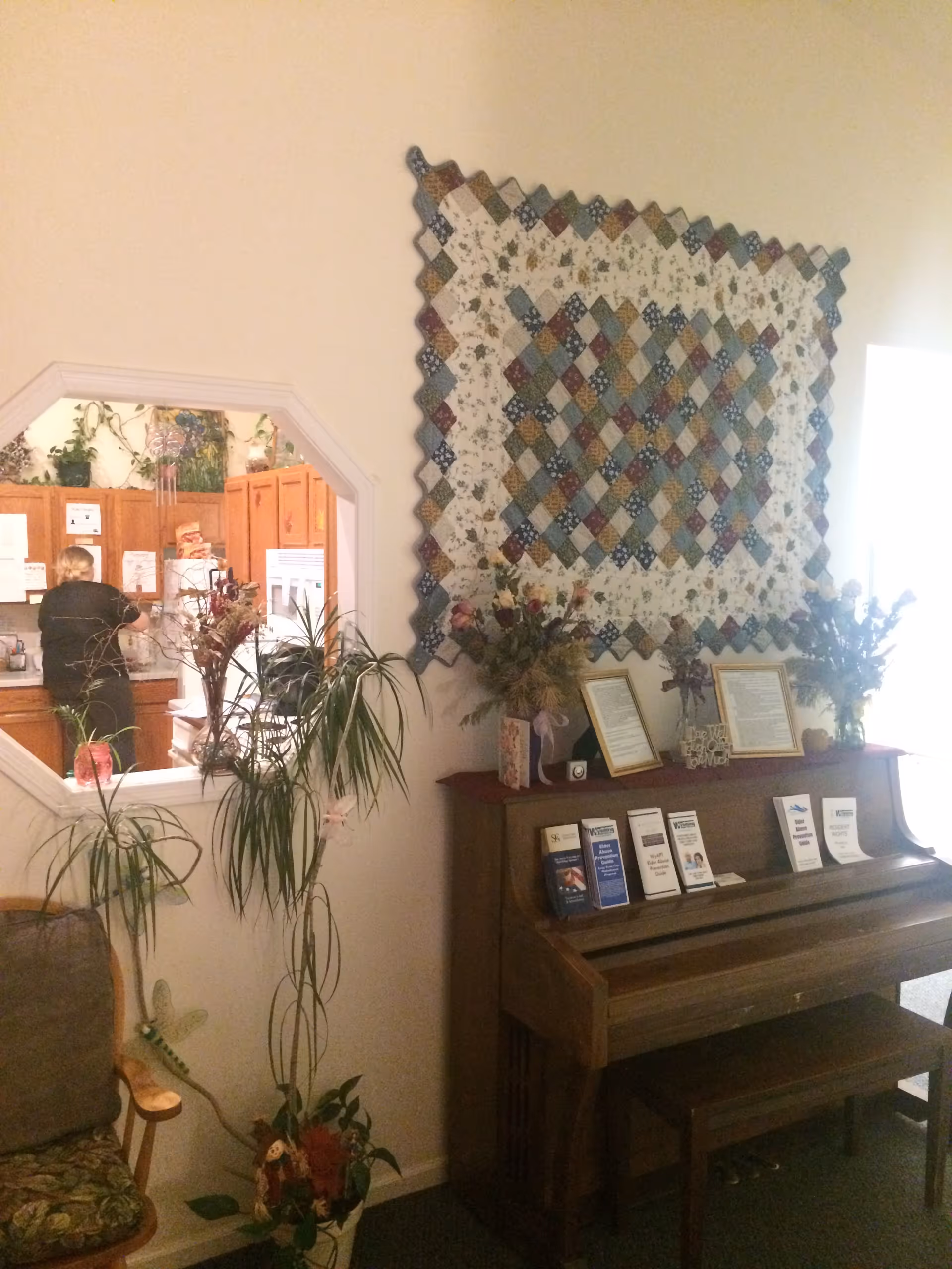 Interior view of a senior living facility showing a wooden piano with brochures and framed documents on top, a colorful quilt hanging on the wall above the piano, a tall potted plant next to the piano, and a partial view into a kitchen area where a person is standing near the counter.