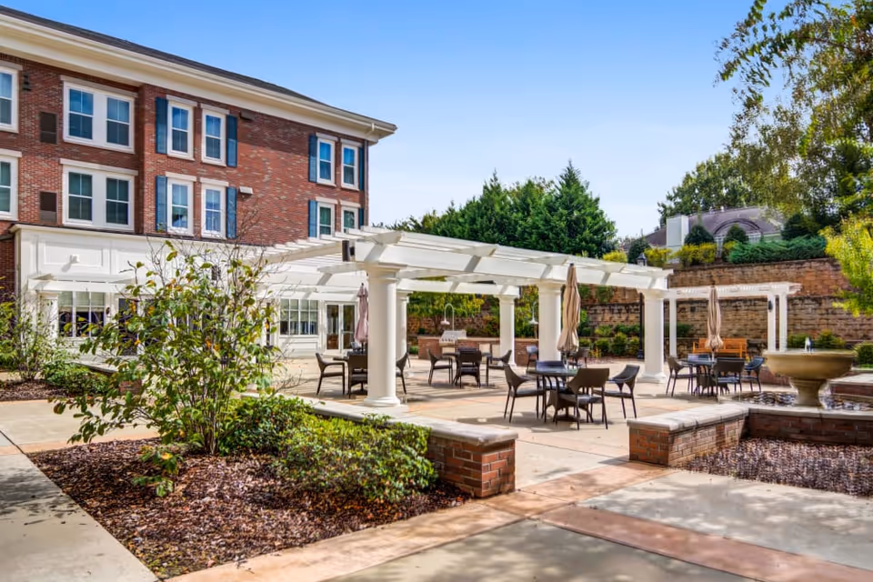 Outdoor patio area at Merrill Gardens at Madison featuring multiple tables with chairs and umbrellas under white pergolas, surrounded by greenery and a brick building in the background.