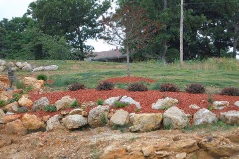 Mulched rock garden with shrubs and boulders in front of a grassy area and trees with a partial roof visible in the background.