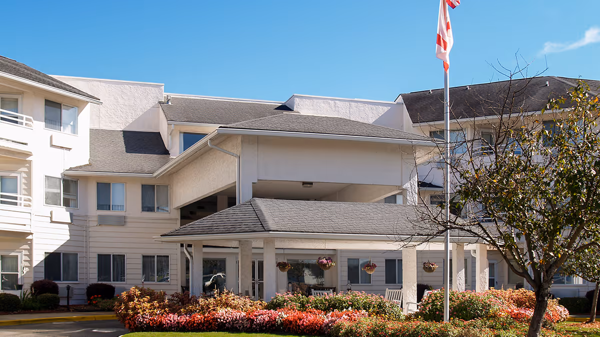 Exterior view of Asher Point Independent Living of Montgomery, showing a multi-story building with white walls and a covered entrance. There are colorful flower beds and a tree in front, with a flagpole displaying a flag. The sky is clear and blue.