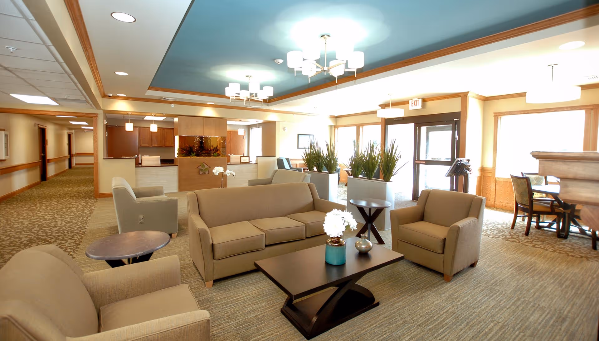 Bright furnished lounge area in a senior living facility with beige sofas and armchairs arranged around a coffee table and a reception desk in the background.