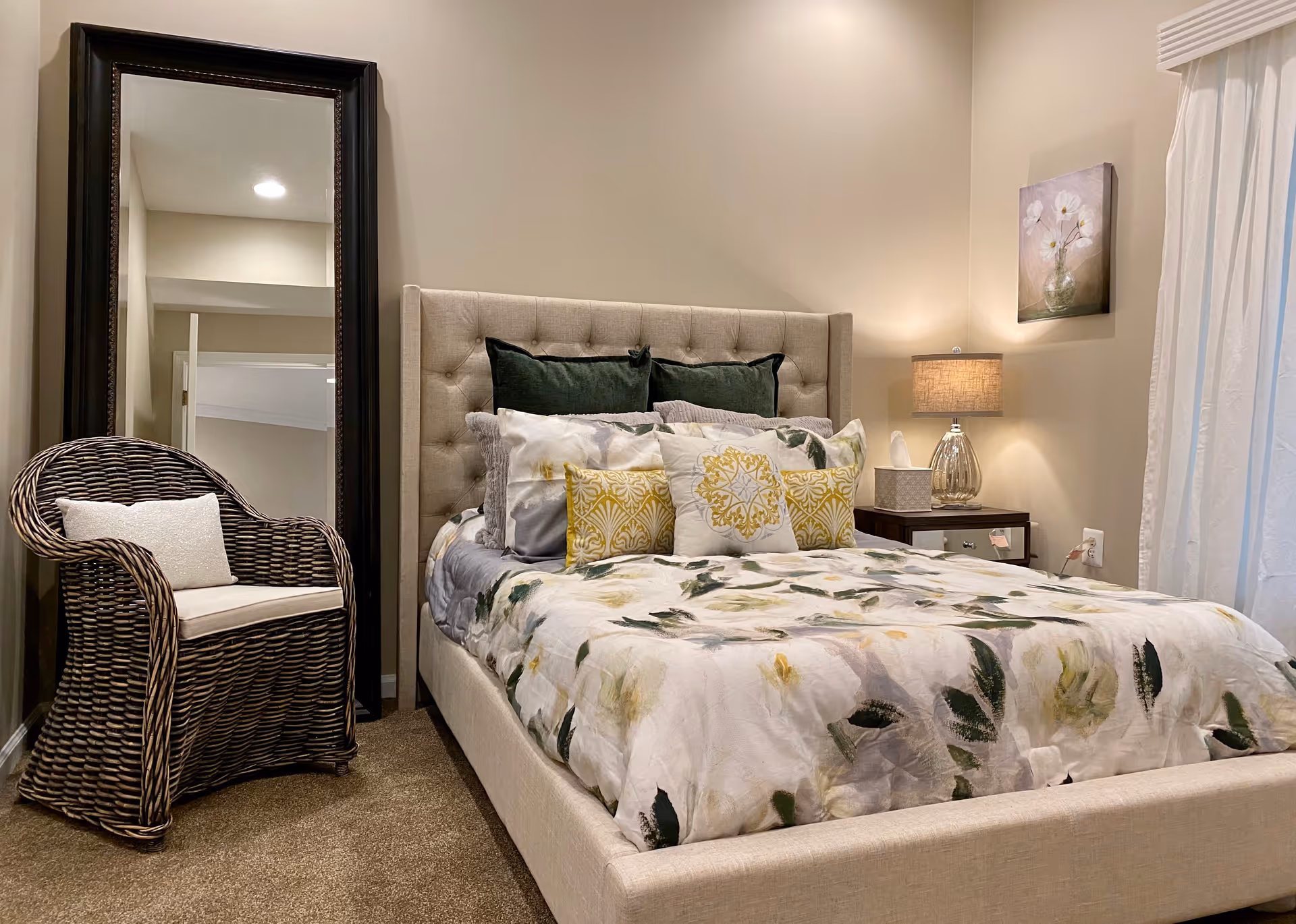 A cozy bedroom with a beige upholstered bed featuring floral bedding and multiple pillows. To the left of the bed is a wicker chair with a white cushion, and behind it stands a tall, dark-framed mirror. On the right side of the bed, there is a nightstand with a lamp, a tissue box, and a small decorative item. A floral painting hangs on the wall above the nightstand, and white curtains cover a window on the far right.