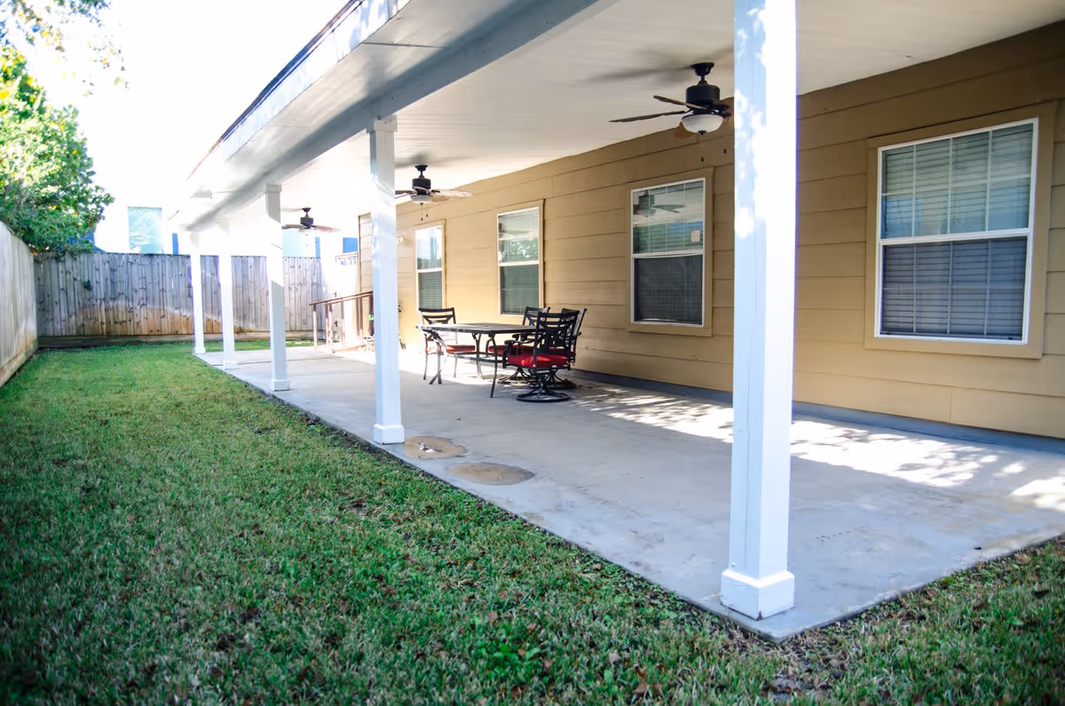 Covered outdoor patio area with a concrete floor, white support columns, ceiling fans, and a table with four chairs with red cushions. The patio is adjacent to a beige building with three windows and is bordered by a grassy lawn and a wooden fence.