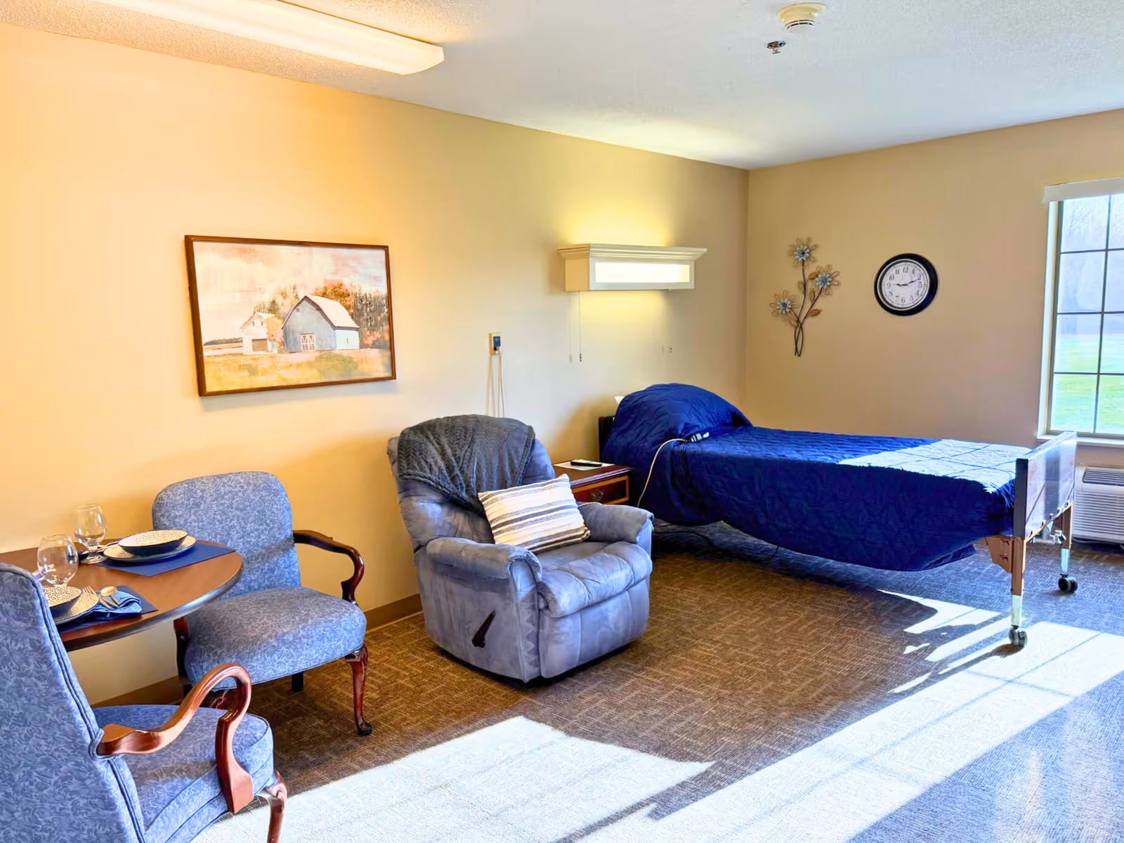 A bright and cozy assisted living bedroom with a single bed covered in a blue quilt, a comfortable recliner chair with a striped pillow, two upholstered chairs around a small round table set with plates and glasses, a wall clock, a window letting in natural light, and a framed painting on the wall.