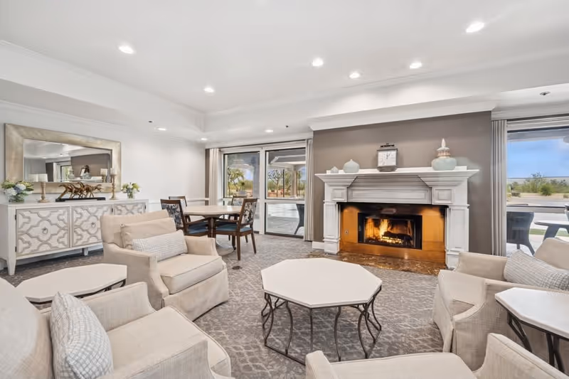 A bright and spacious living room with beige armchairs arranged around a white octagonal coffee table. There is a white fireplace with a fire burning and decorative items on the mantel. Behind the seating area, there is a dining table with chairs near large sliding glass doors that open to an outdoor patio. The room has a neutral color palette with light walls, a patterned carpet, and recessed ceiling lights.