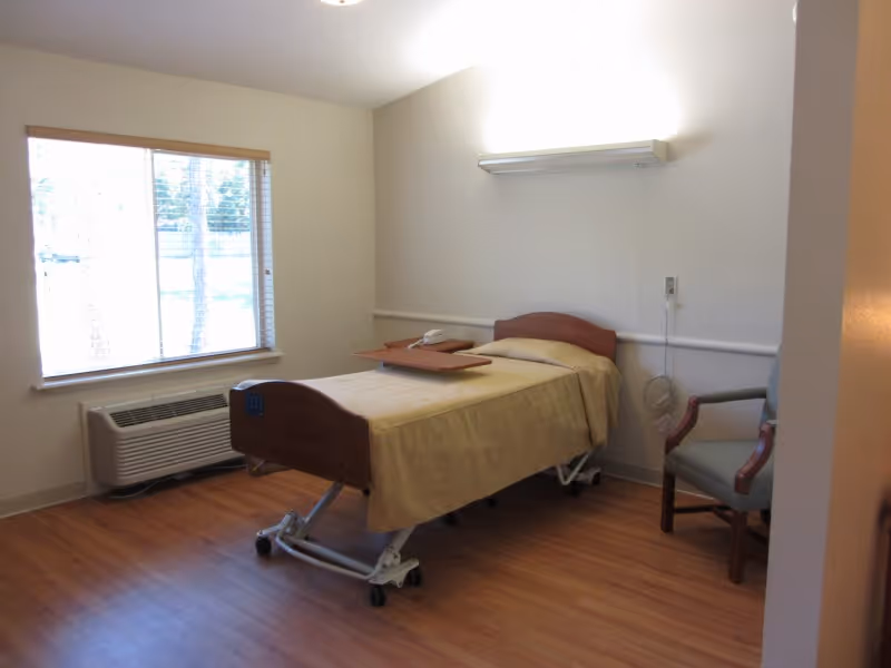 A simple healthcare facility room with a single hospital bed covered with a beige blanket, a wooden overbed table, a gray armchair, a large window with blinds, and a wall-mounted air conditioning unit below the window. The room has light-colored walls and wood flooring.