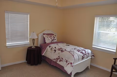 A small bedroom with beige walls and carpeted floor featuring a single white bed with floral bedding in shades of purple and white. There is a round side table with a lamp next to the bed, and two windows with white blinds on adjacent walls.