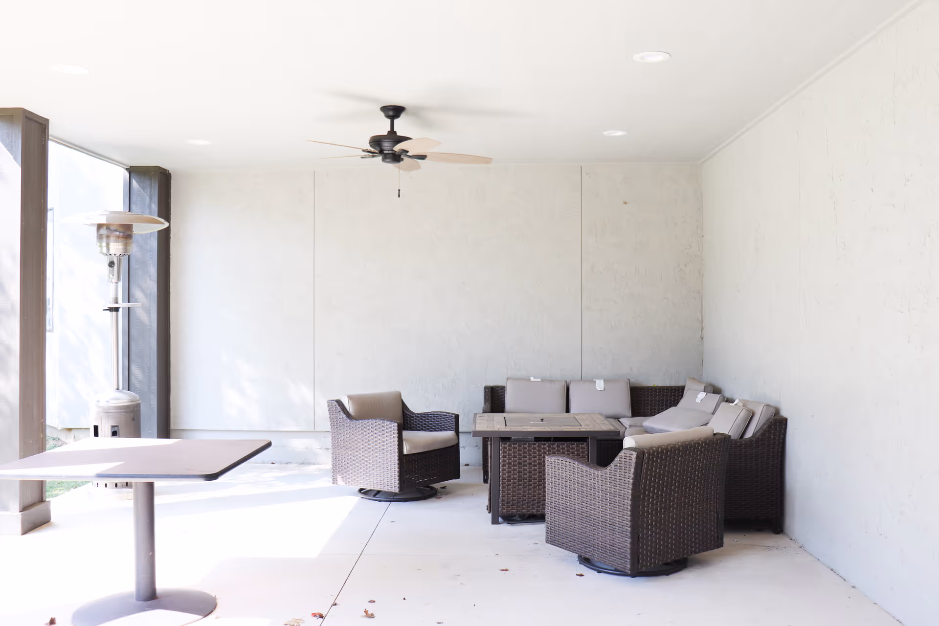 Covered outdoor patio area with a ceiling fan, a patio heater, a table, and a set of cushioned wicker chairs arranged around a square table against a light-colored wall.