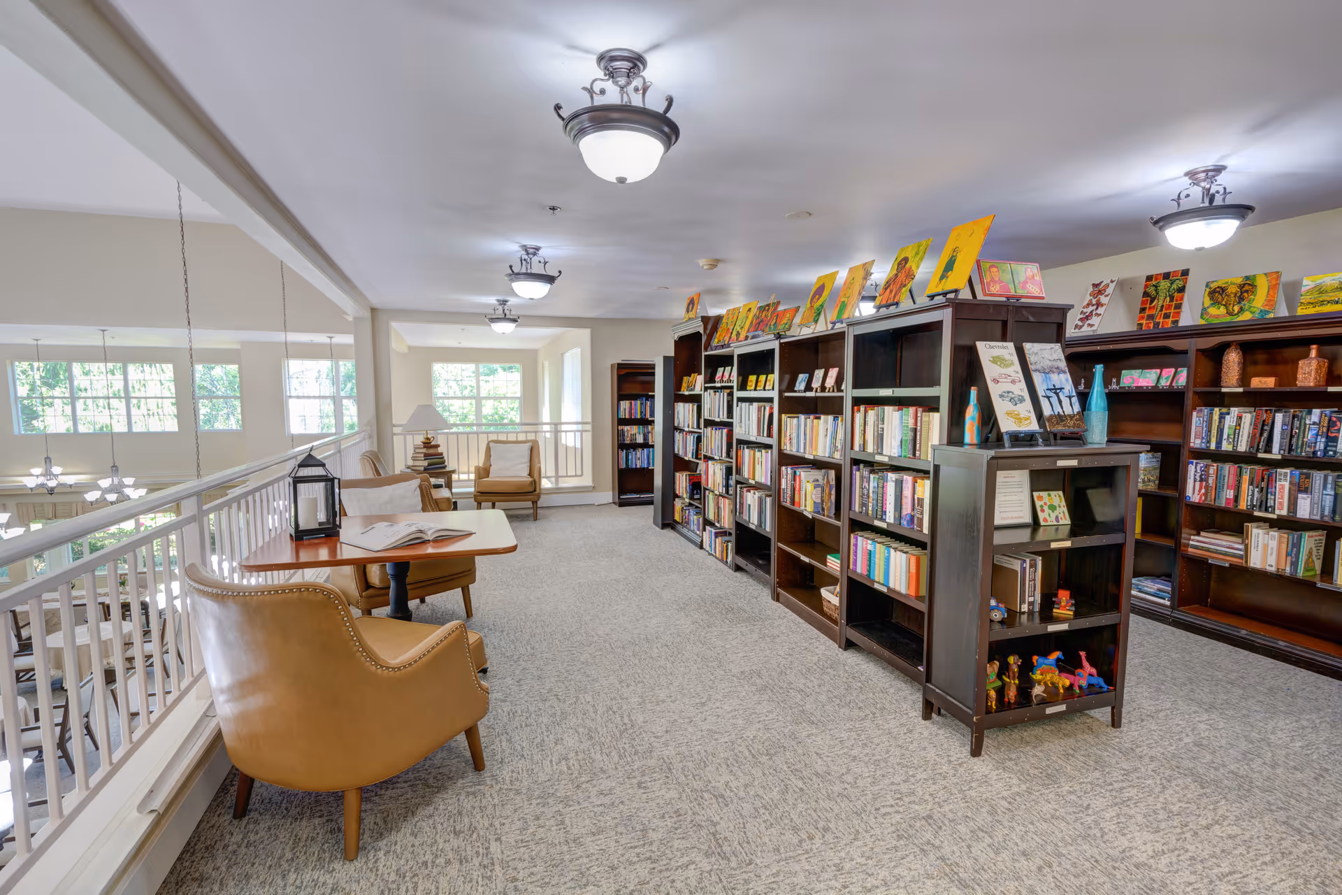 A bright and spacious library area in Redwood Heights by Cogir featuring multiple dark wood bookshelves filled with books and decorative items. There are several colorful paintings displayed on top of the shelves. Comfortable tan leather chairs and small tables are arranged near a white railing overlooking a lower level with large windows letting in natural light.