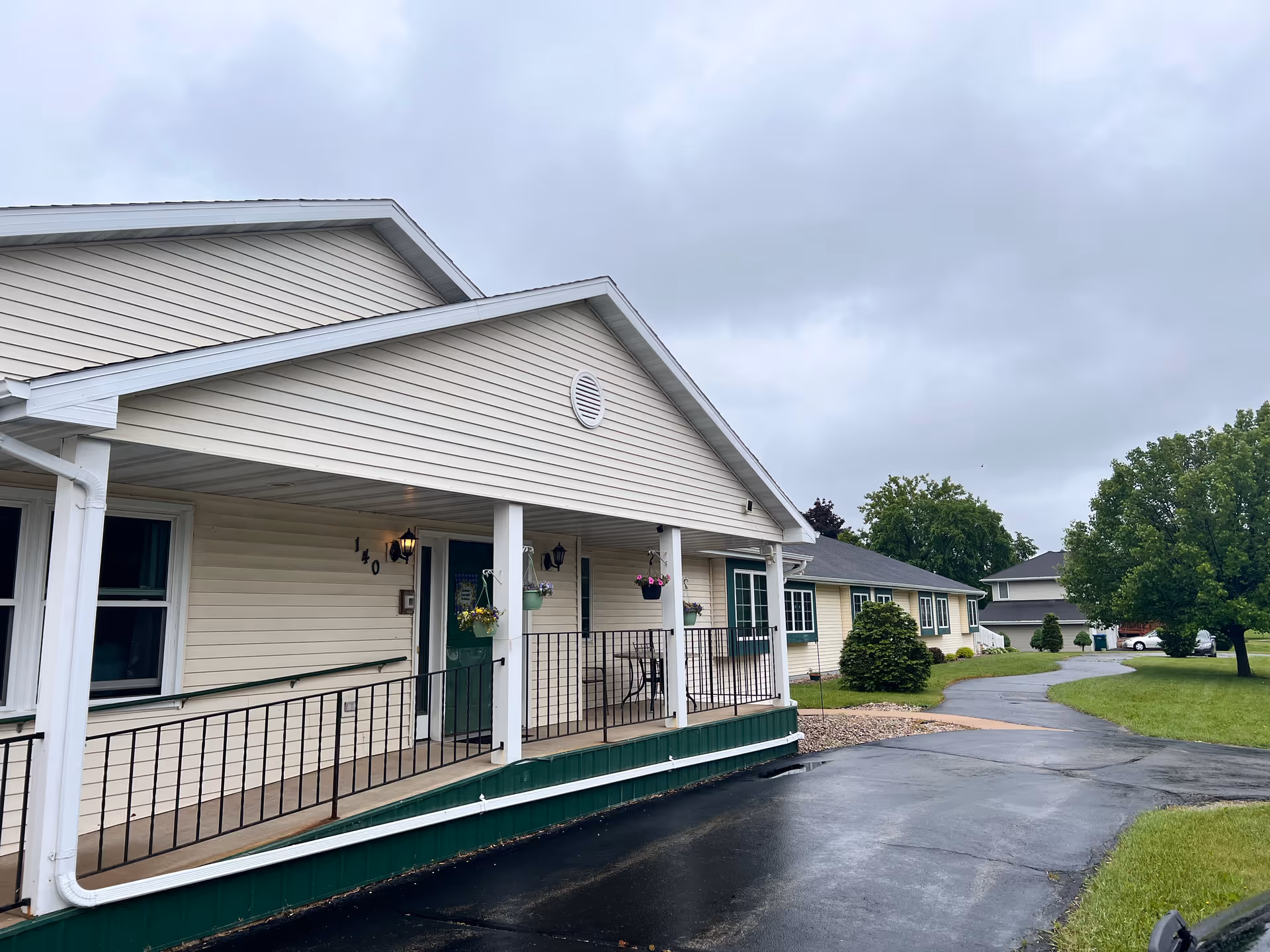 Covered front porch and entrance of a single-story assisted living building with a ramp, driveway, and lawn under a cloudy sky.